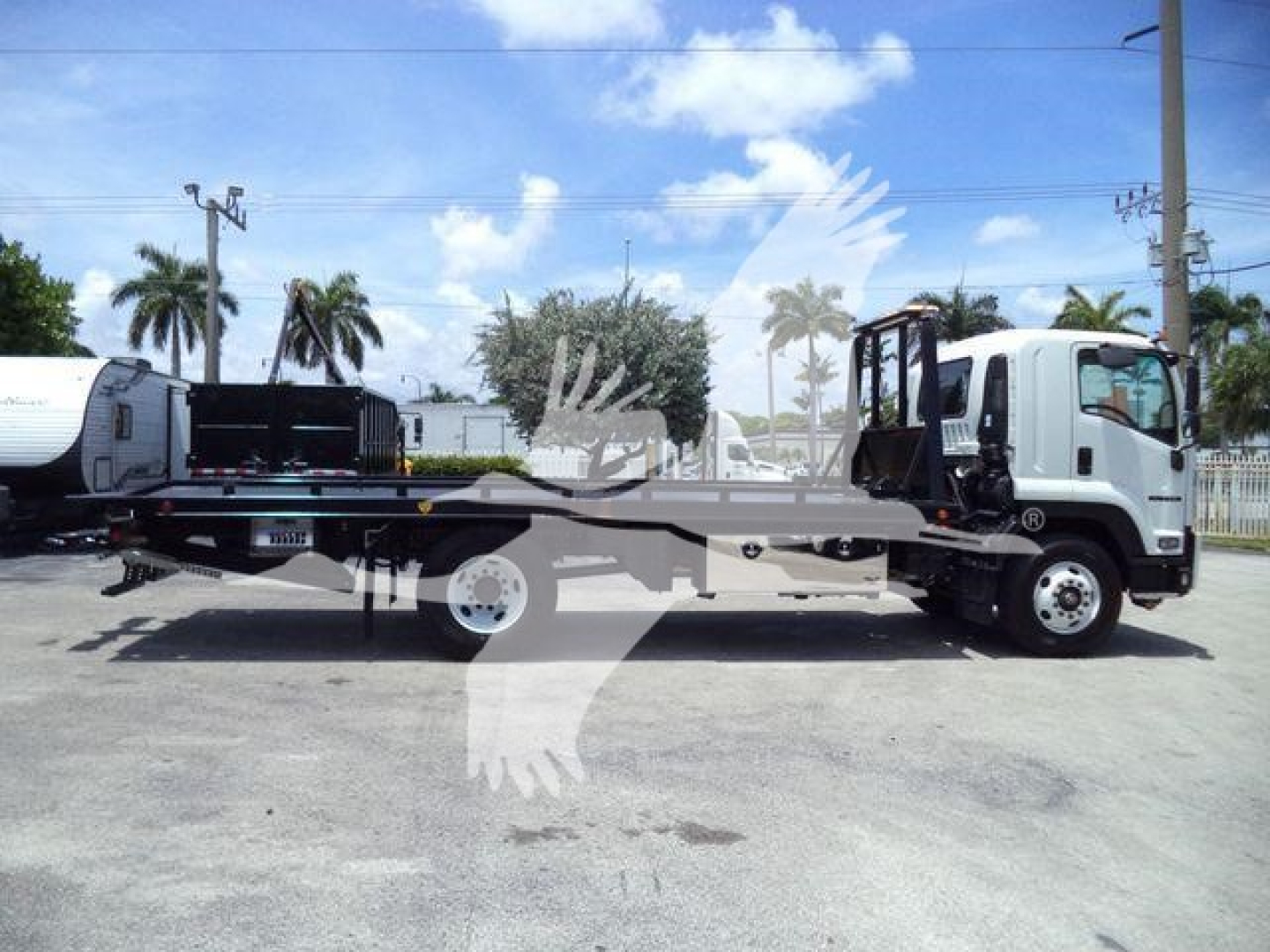Truck bed tow truck with Isuzu flatbed chassis and hydraulic equipment, side view. Displayed stock and dealership lot.