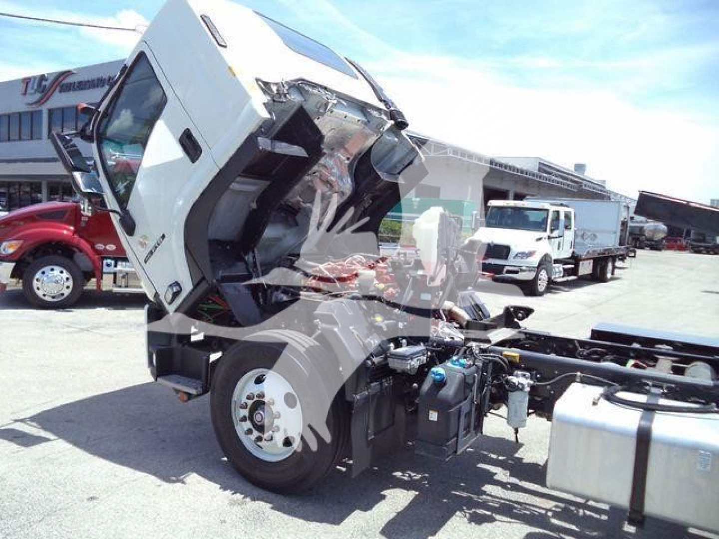 Isuzu FTR truck cab lifted open showing engine bay with fuel tank and chassis; front wheel and frame visible; other trucks in background.