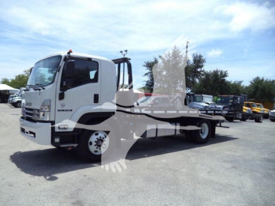 Isuzu FTR tow truck chassis with flatbed, white cab, side view showing wheel, clear windshield, and equipment yard in background.