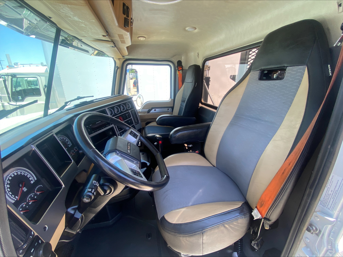 Mack PI64T truck cab interior showing steering wheel dashboard and two-tone seat upholstery with seat belt. Dash gauges visible.