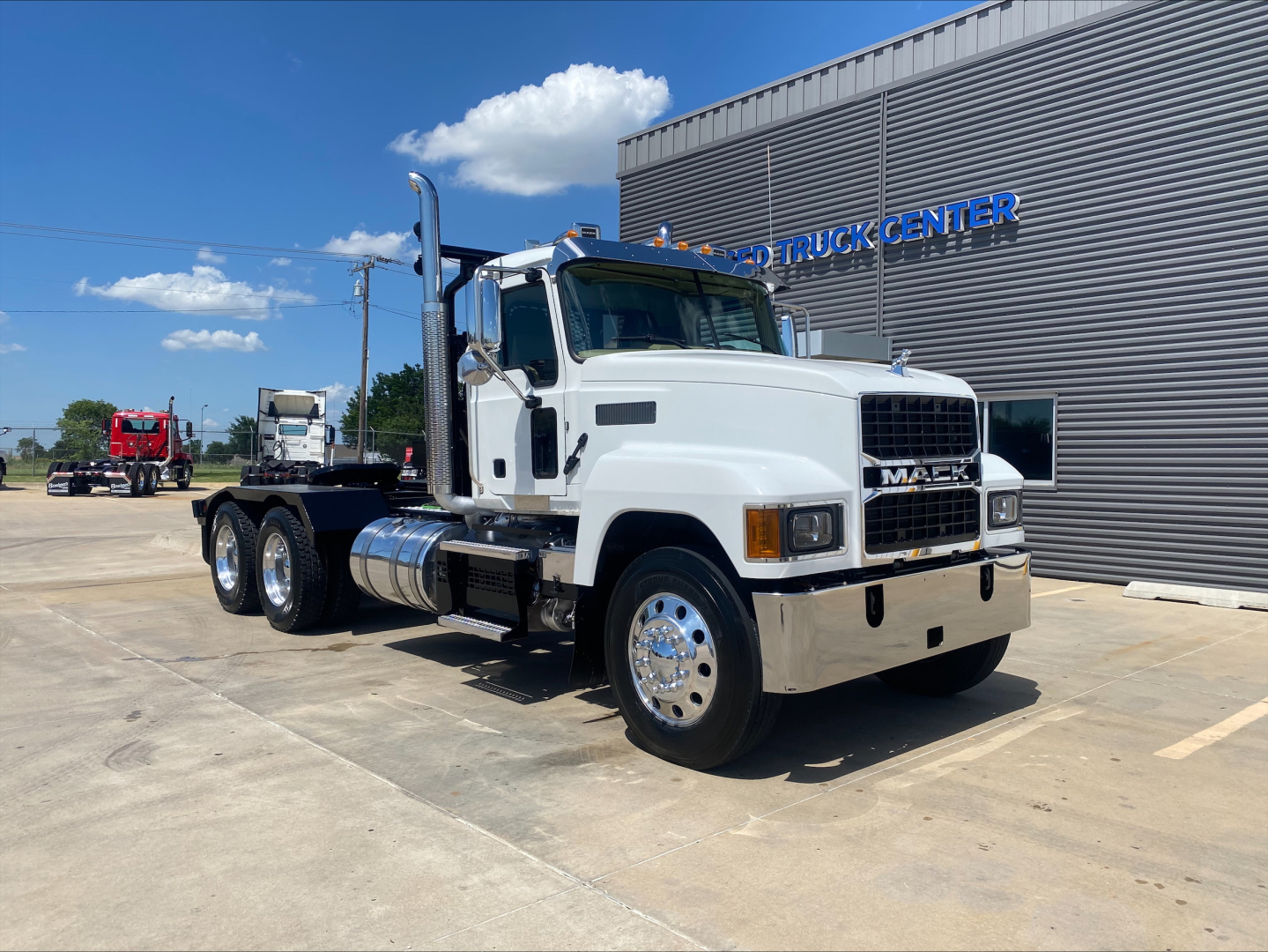 2024 Mack PI64T heavy truck with winch, white cab, chrome fuel tanks, and dual rear axles in front of a building. Visible text includes 'MACK' on the grille and 'TRUCK CENTER' on the building.