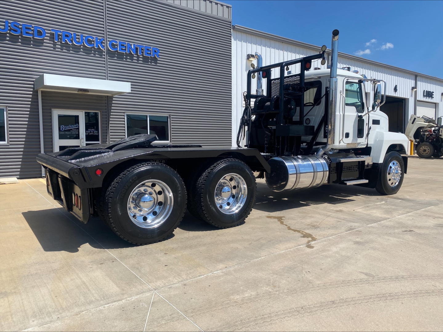 2024 Mack PI64T heavy-duty truck with flatbed and dual rear wheels, chrome fuel tanks, and rear winch setup visible at dealership.
