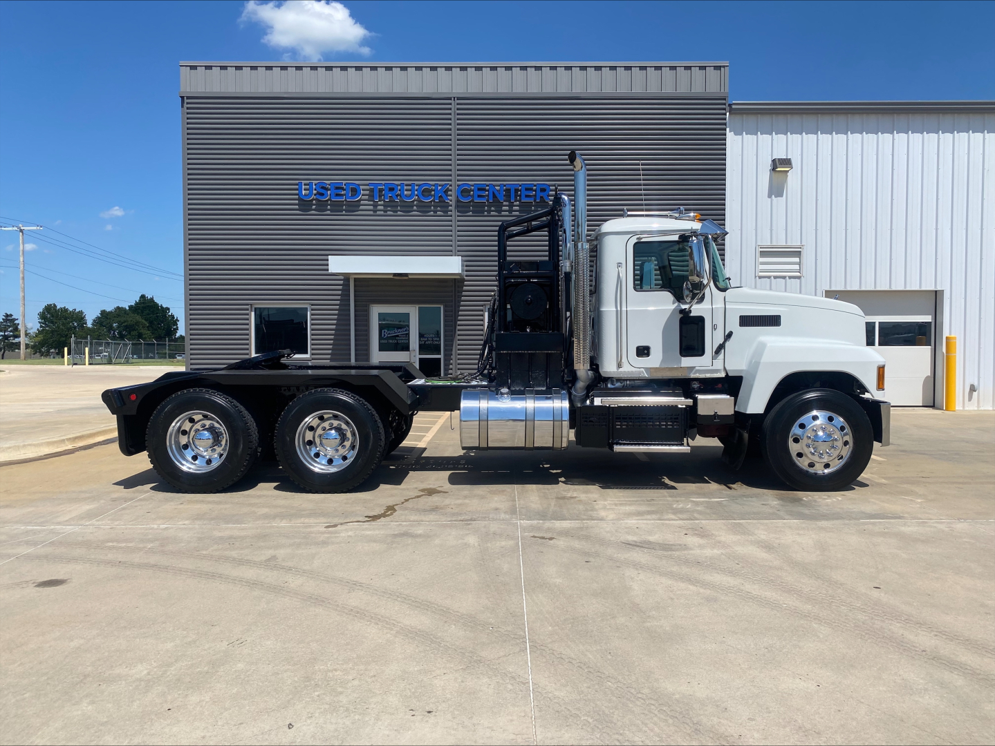 Truck side view showing a white 2024 Mack PI64T utility tractor with dual rear axles and a winch, parked in front of a building marked USED TRUCK CENTER.