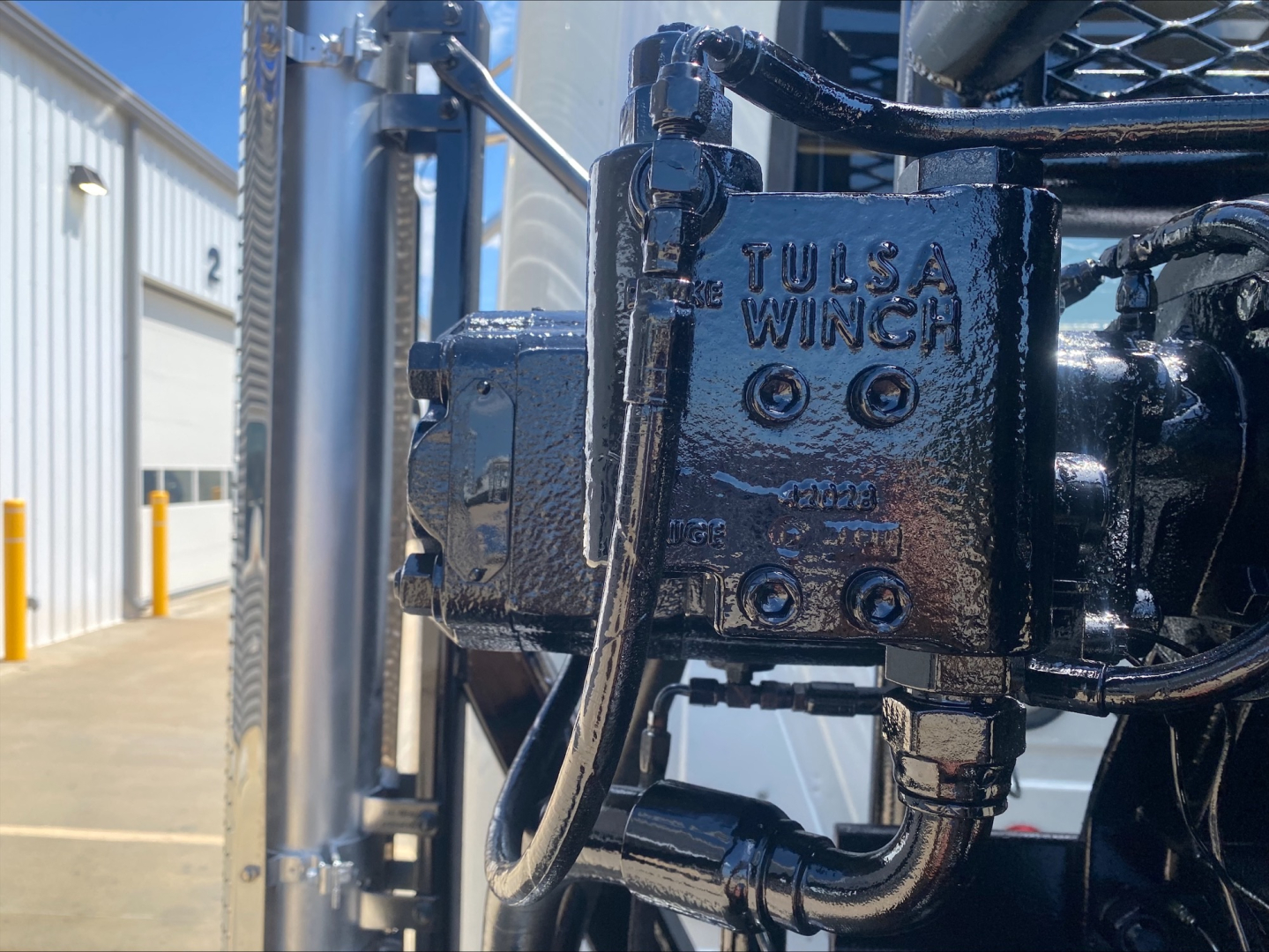 Winch component labeled Tulsa Winch with hydraulic hoses and mounting on Mack PI64T truck, close-up view.