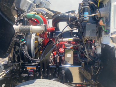Engine bay of a Mack PI64T truck showing hoses, hydraulic lines, and components with labels visible.