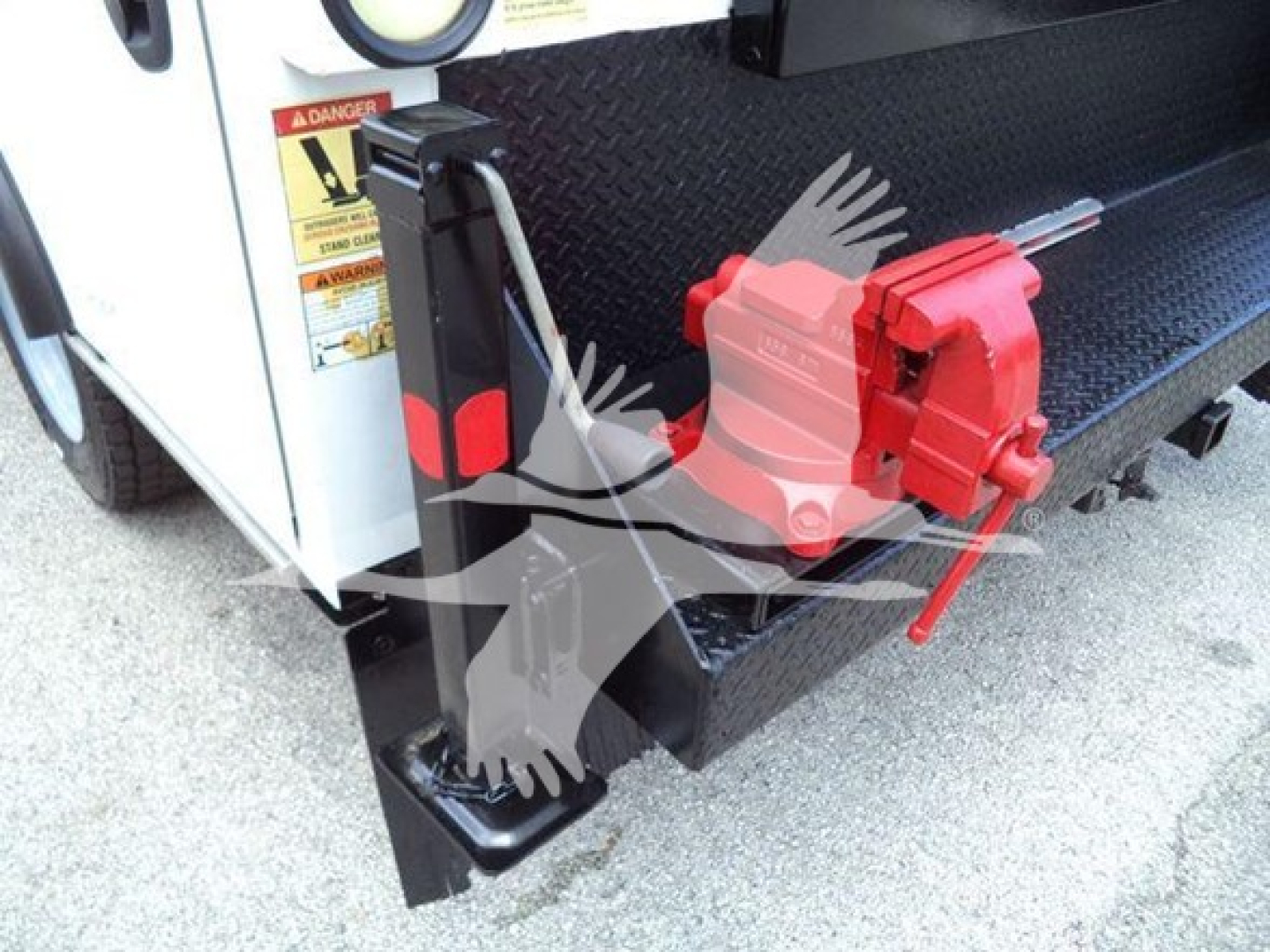 Truck hitch area with black steel platform, red hydraulic jack/vice and warning label. Visible text includes a red warning sticker and a small yellow label; full readable text not clear.