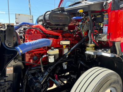 Kenworth T680 engine bay showing red Cummins-style engine with blue radiator hoses, air intake, various hoses and wiring, front wheel and suspension visible.