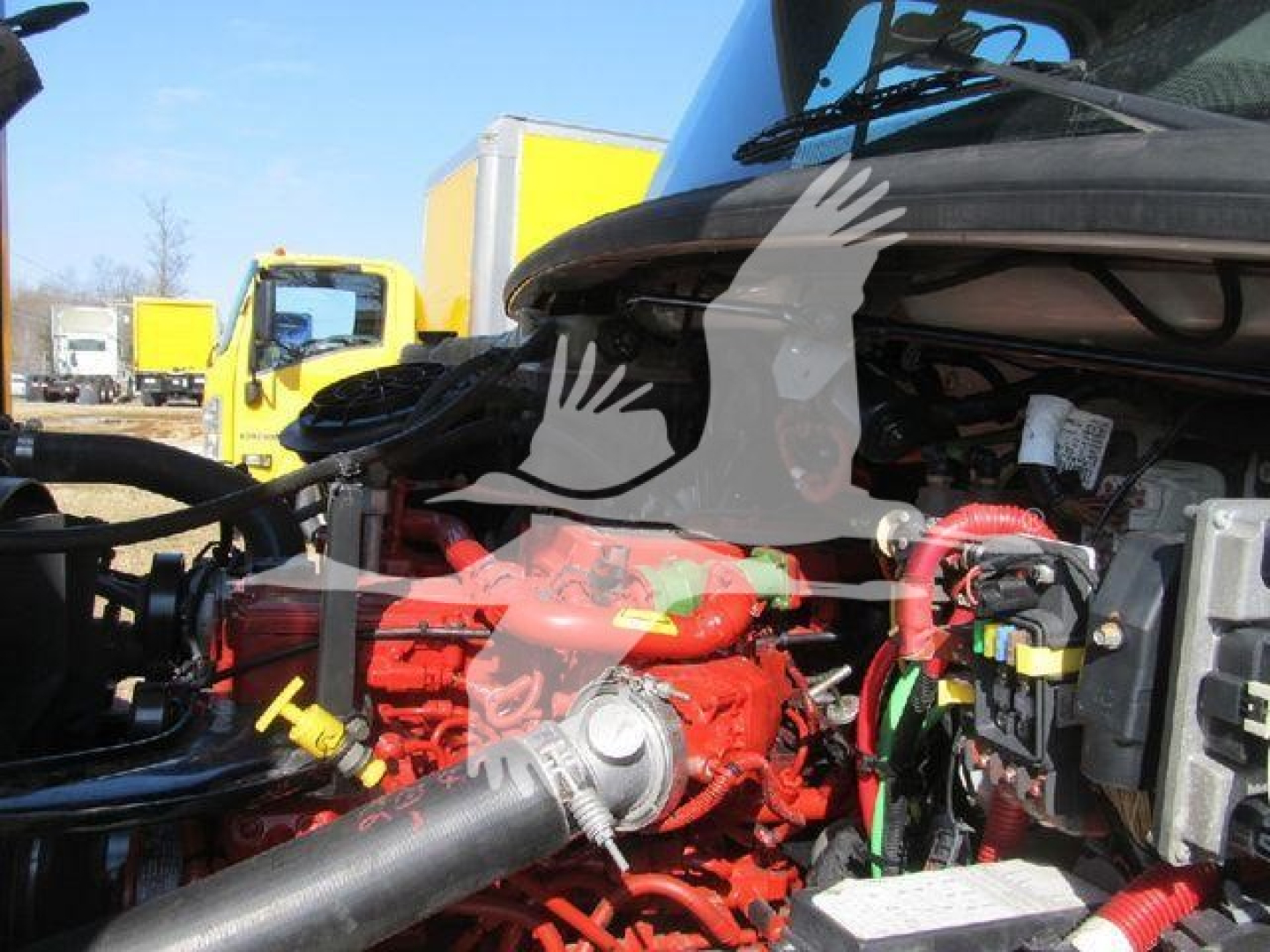 Truck engine bay of a 2019 Freightliner M2 106 with red engine components and various hoses; background shows yellow trucks.