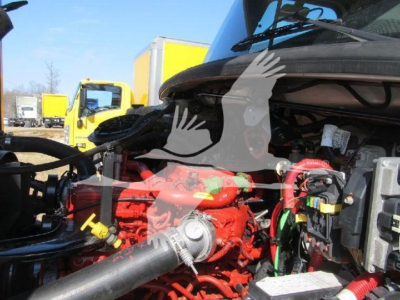 Truck engine bay of a 2019 Freightliner M2 106 with red engine components and various hoses; background shows yellow trucks.