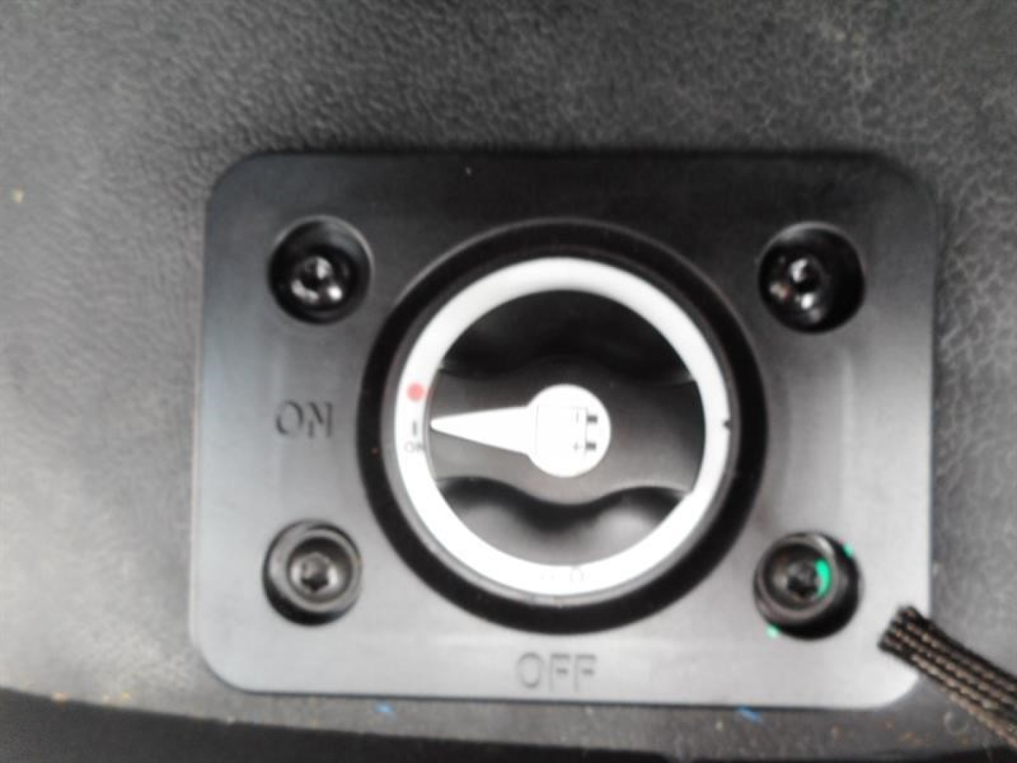 Engine bay switch panel with ON/OFF dial and surrounding screws; circular gauge in center; small green indicator light bottom-right.
