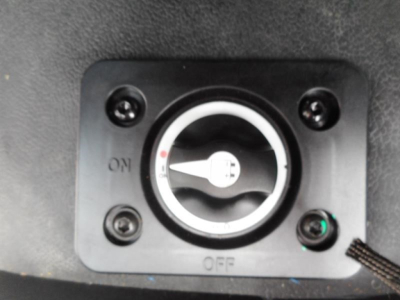 Engine bay switch panel with ON/OFF dial and surrounding screws; circular gauge in center; small green indicator light bottom-right.