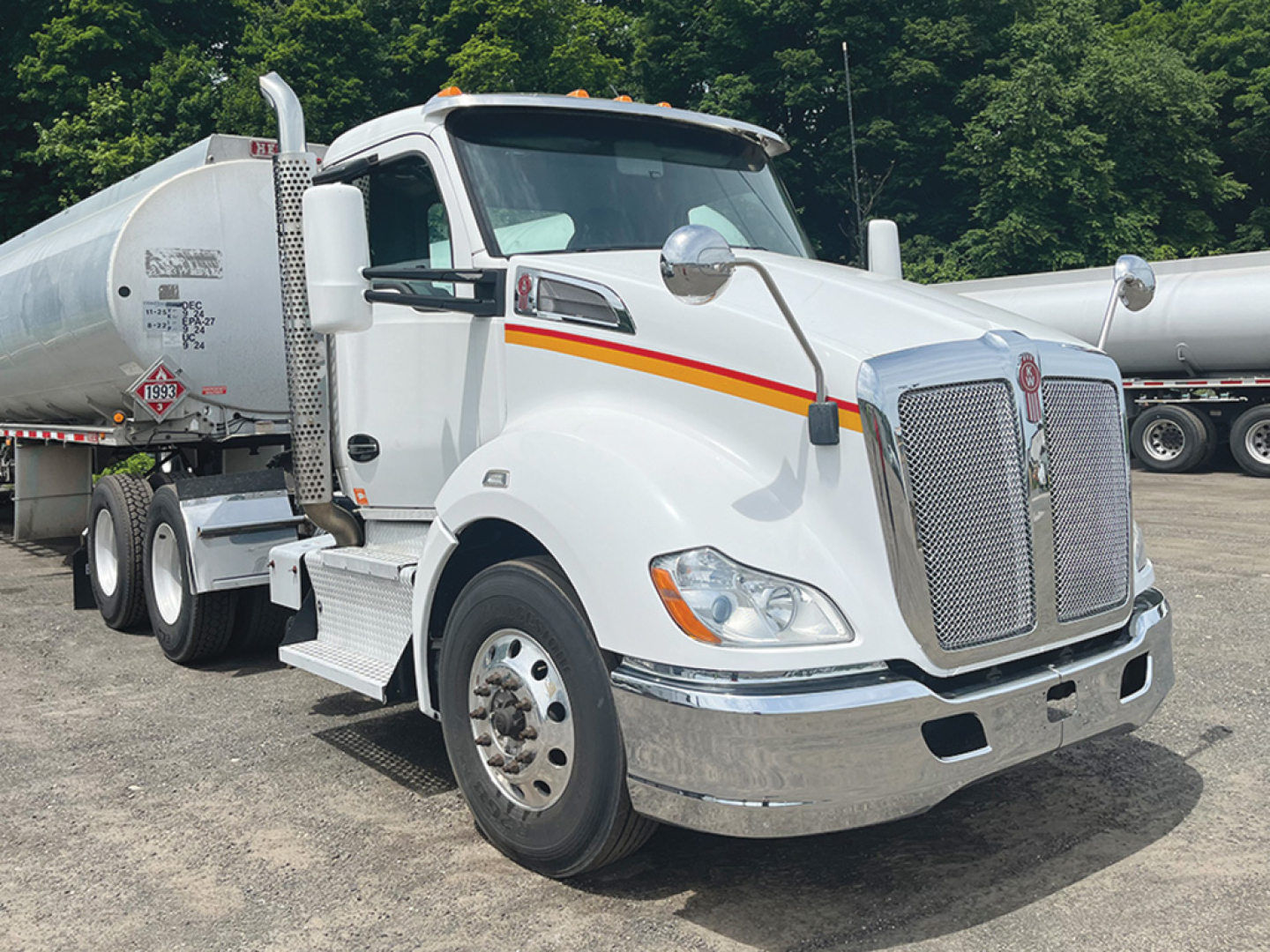 Kenworth T680 conventional daycab truck shown in a parking lot with fuel or liquid tank trailer in background.