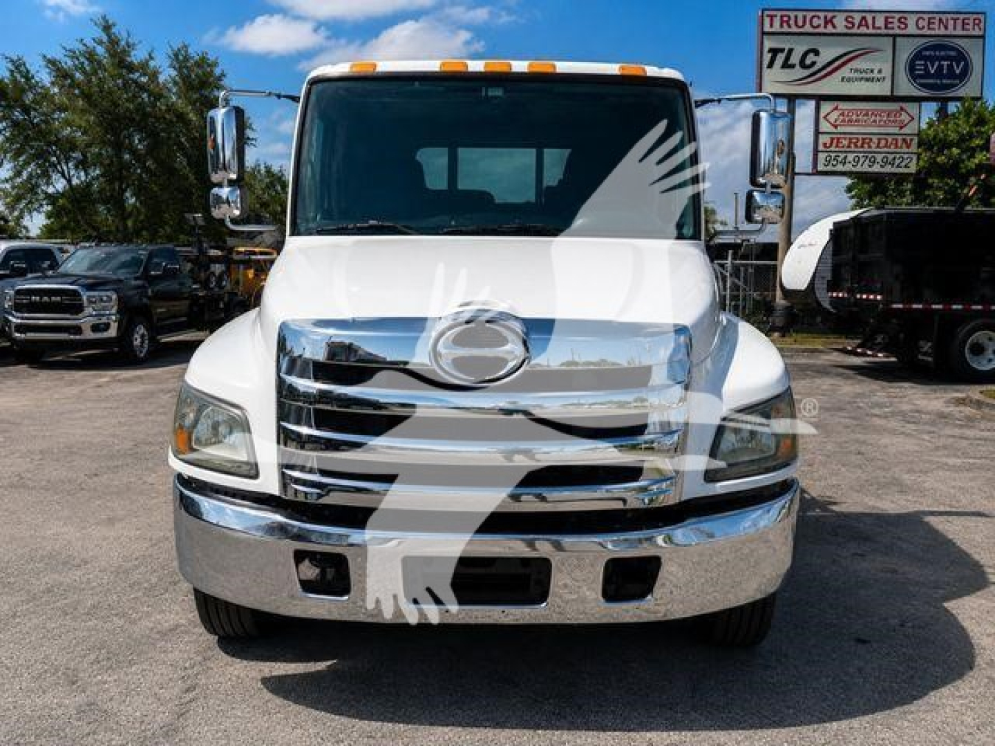 Front view of a white 2020 Hino 258Al tow truck with chrome grille, bumper, and amber clearance lights on the roof, dealership lot in background.