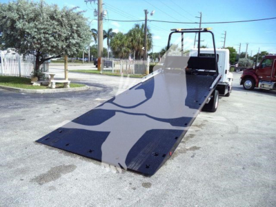Truck rollback with a lowered flatbed ramp and wheel chocks, aluminum surface shows loading deck, rear wheels, and operator cab in background.