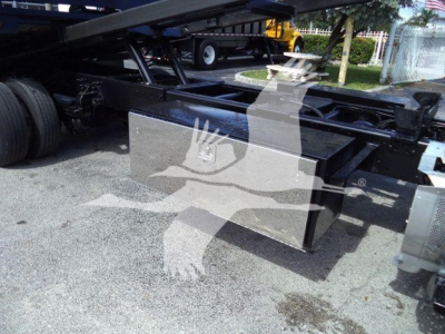 Truck rollback chassis with a rectangular metal toolbox mounted beneath the frame showing a reflective stainless surface and hydraulic components.