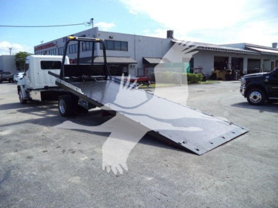 Wheel-lift rollback truck with a metal incline ramp on a concrete lot, Hino 258Al cargo/tow truck in background, dealership building in distance