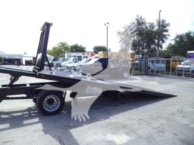 Tow truck tilt-bed with hydraulic rollback in yard; visible text on sticker and controls faintly legible.