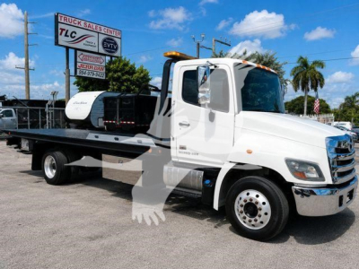 Hino 258Alp rollback tow truck in a dealership lot, white cab, flatbed with equipment, sign reading TLC Truck Sales Center in background.
