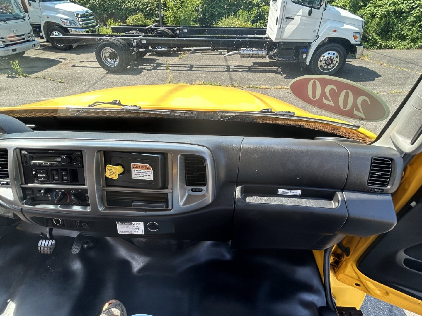 Dashboard and interior view of a yellow 2020 Hino 338 box van from the passenger side, with a stickered 2020 tag on the windshield, and a view of the exterior cab and chassis.