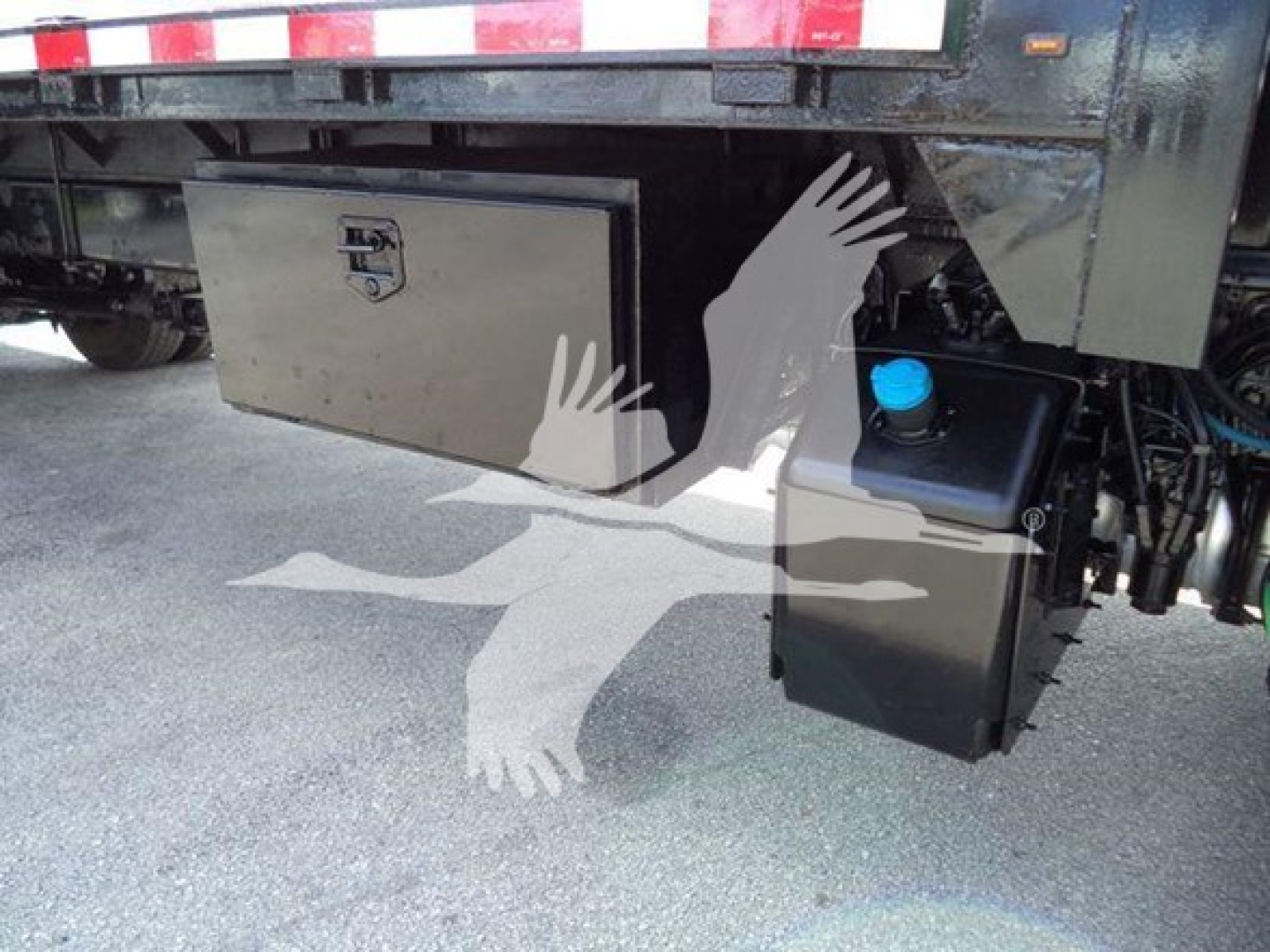 Flatbed truck undercarriage showing a metal toolbox and black plastic reservoir mounted near the rear, with a blue cap visible; reflective tape on the trailer above.