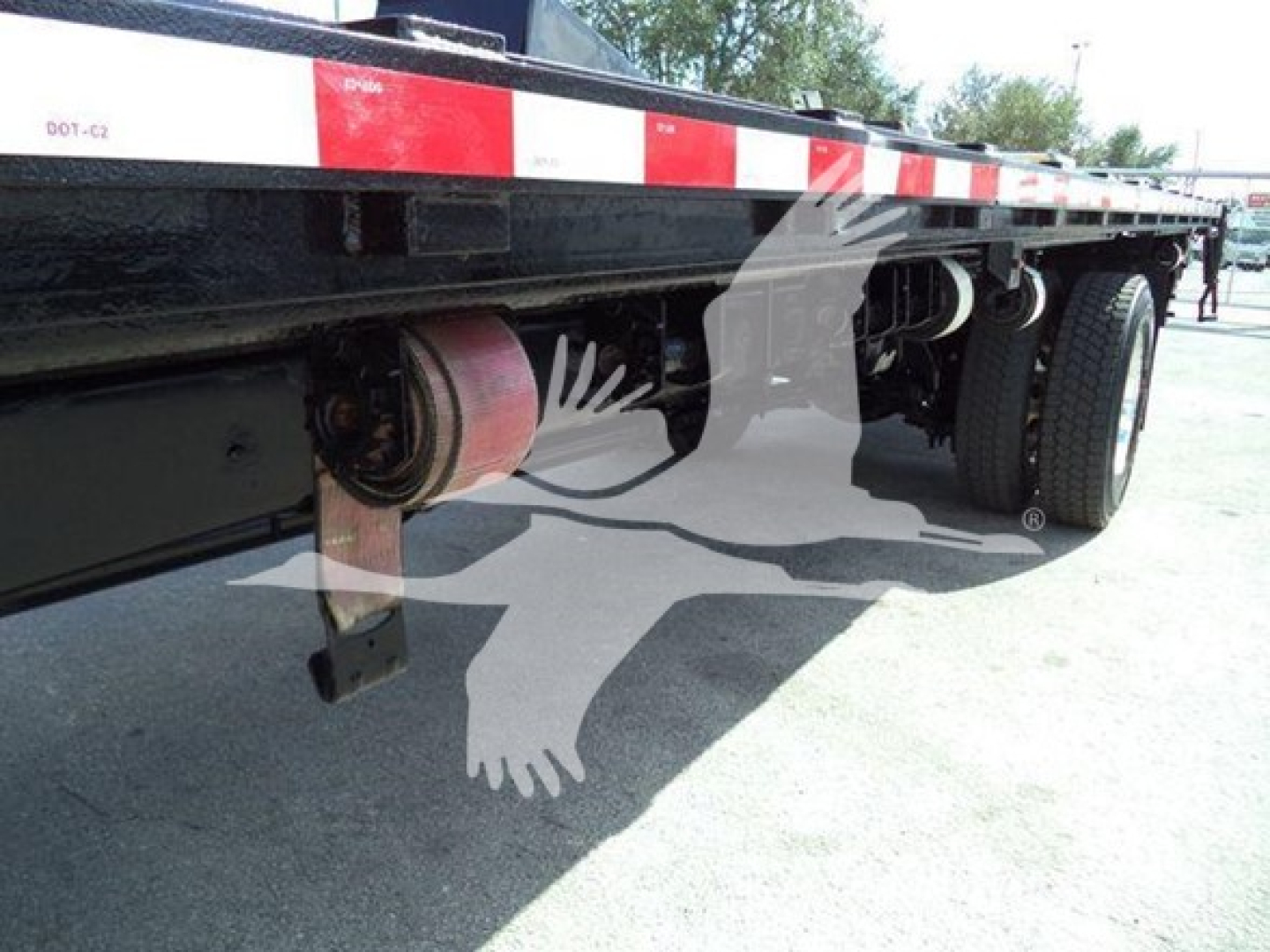 Flatbed semi-trailer undercarriage showing suspension and wheel assembly with cylindrical components, chassis beams, and landing gear in view.