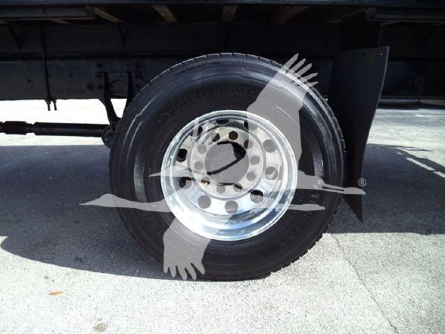 Flatbed truck wheel and tire under chassis; steel rim with lug nut holes visible, undercarriage and suspension components