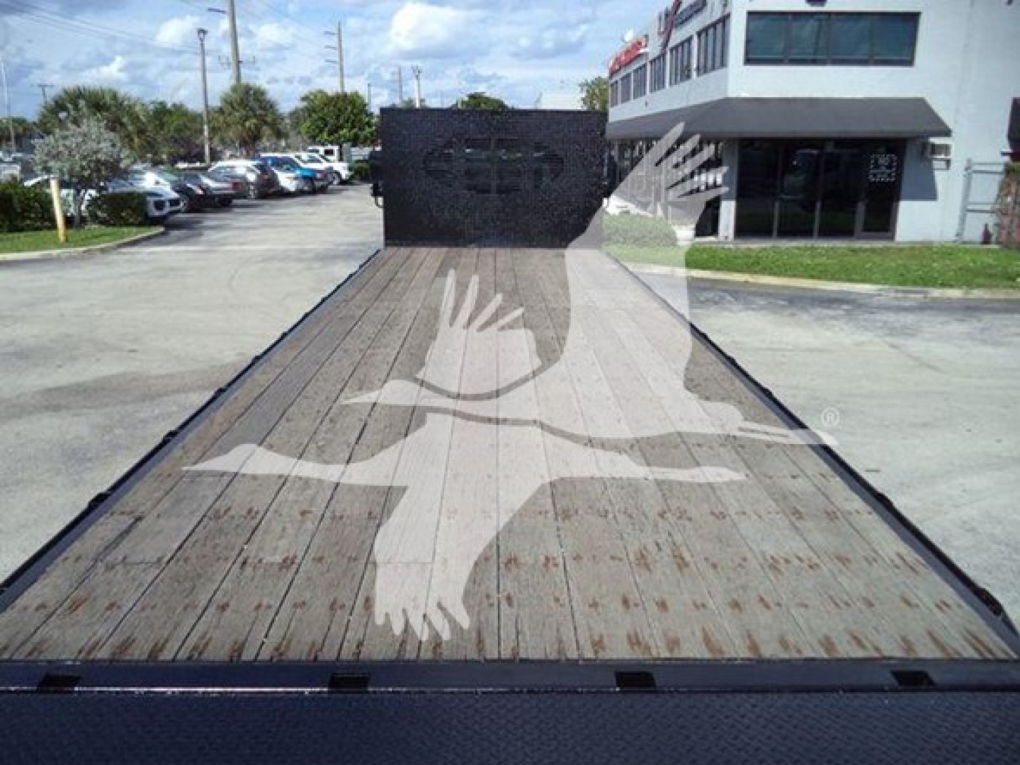 Flatbed truck bed showing wooden deck and a tall black cab shield in background; International logo watermark overlayed; generic dealership lot in distance.