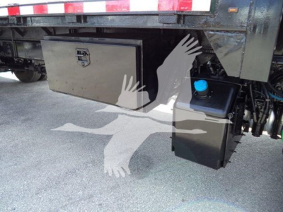 Flatbed truck undercarriage showing a metal toolbox and black plastic reservoir mounted near the rear, with a blue cap visible; reflective tape on the trailer above.