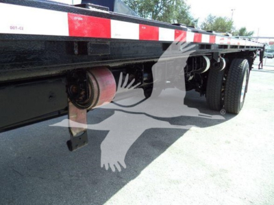 Flatbed semi-trailer undercarriage showing suspension and wheel assembly with cylindrical components, chassis beams, and landing gear in view.