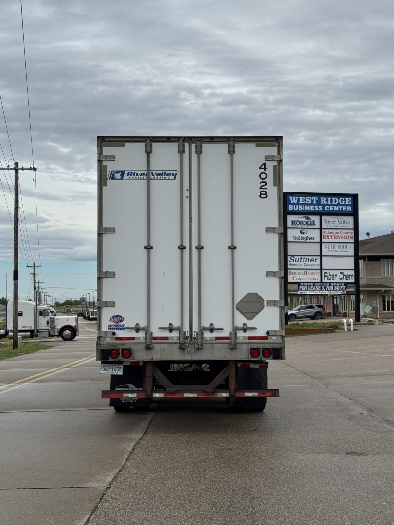 Rear view of a white utility trailer with double doors, brand stickers, and taillights showing in a parking lot.