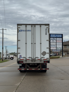 Rear view of a white utility trailer with double doors, brand stickers, and taillights showing in a parking lot.