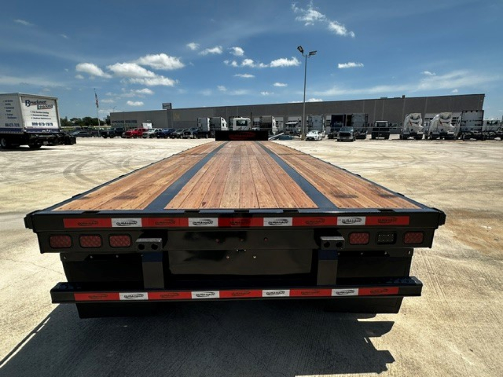 Flatbed trailer deck showing wood planks with black steel framework and rear LED taillights; a truck yard with buildings in background.