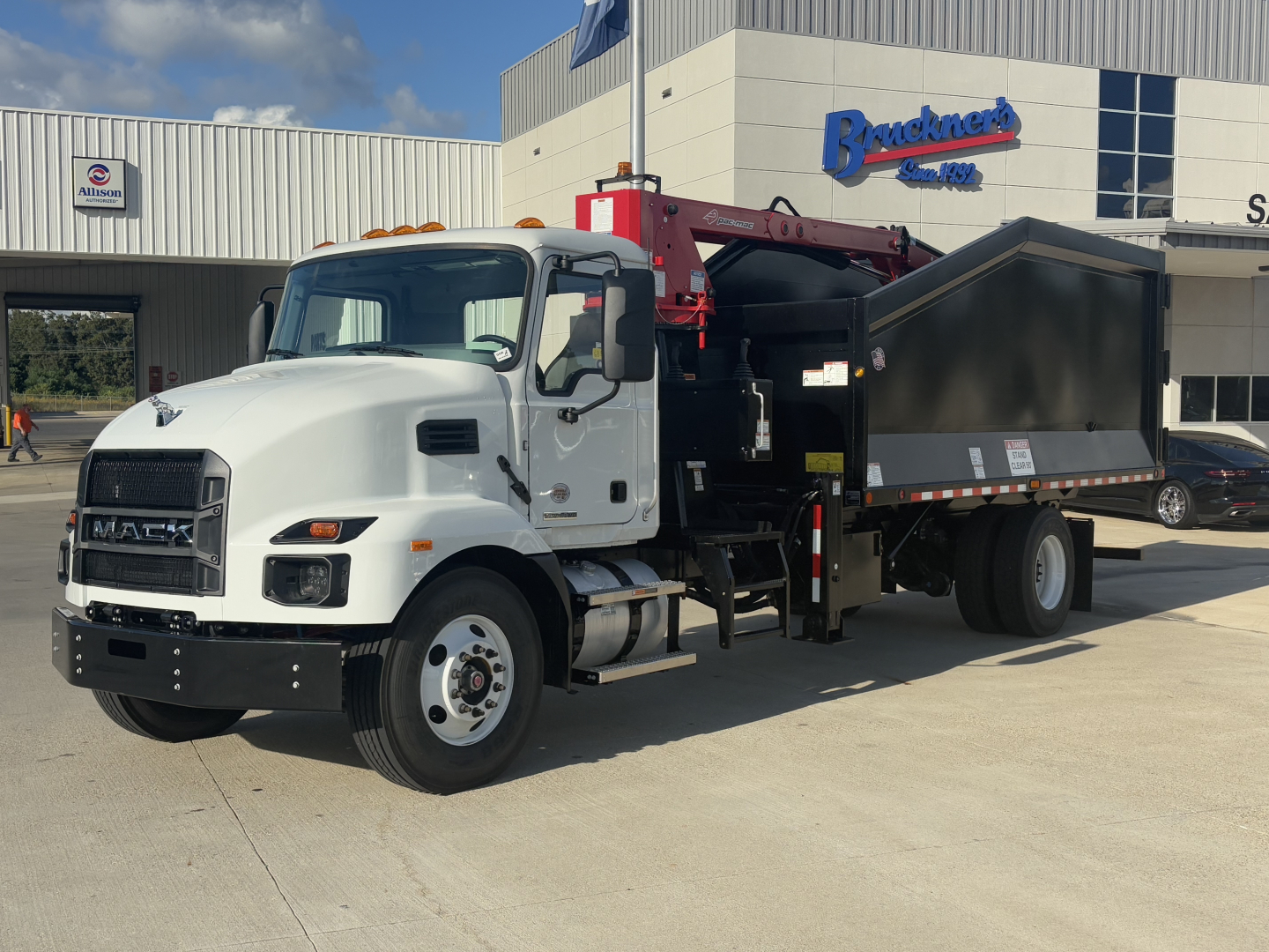 2026 Mack MD7 grapple truck shown in a dealership lot with a white cab and red crane arm, black dump hopper, and Bracken's signage in background.