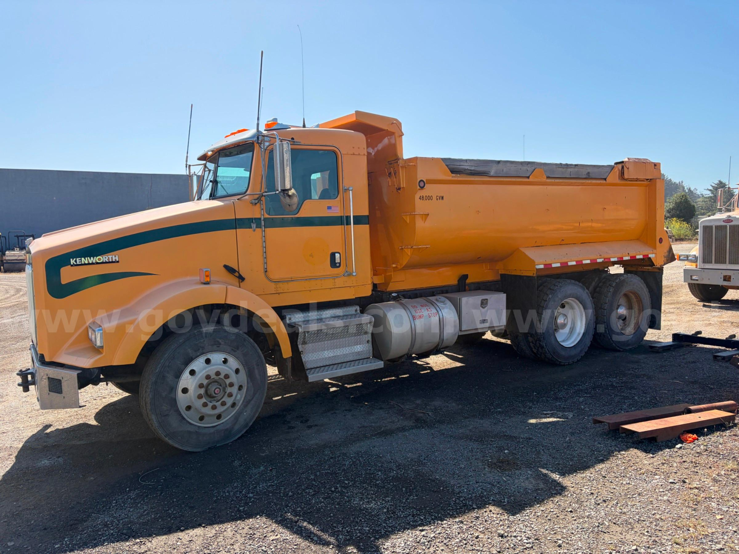Kenworth T800B dump truck in service showing orange cab and dump bed with fuel tanks and wheels visible.