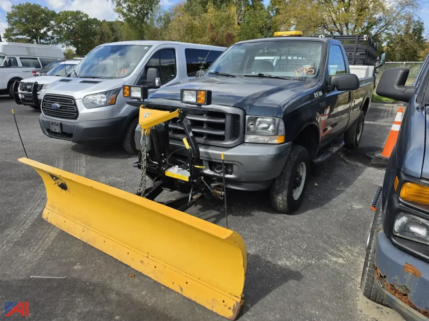 Ford F-250 Super Duty pickup with front plow and liftgate attachment in a parking lot; blue-gray crew cab truck with yellow snow plow blade, plow mounting hardware, and orange construction cone nearby.