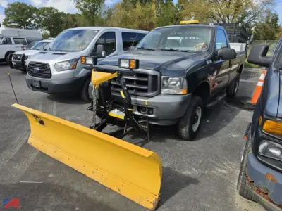 Ford F-250 Super Duty pickup with front plow and liftgate attachment in a parking lot; blue-gray crew cab truck with yellow snow plow blade, plow mounting hardware, and orange construction cone nearby.
