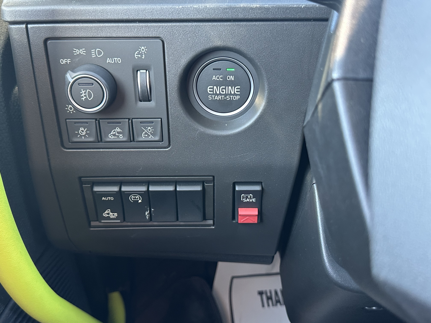 Truck dash area showing engine start-stop button and control switches around left side; circular engine start-stop with ACC ON text; headlight and fog light controls; various toggles and a red hazard-like switch.