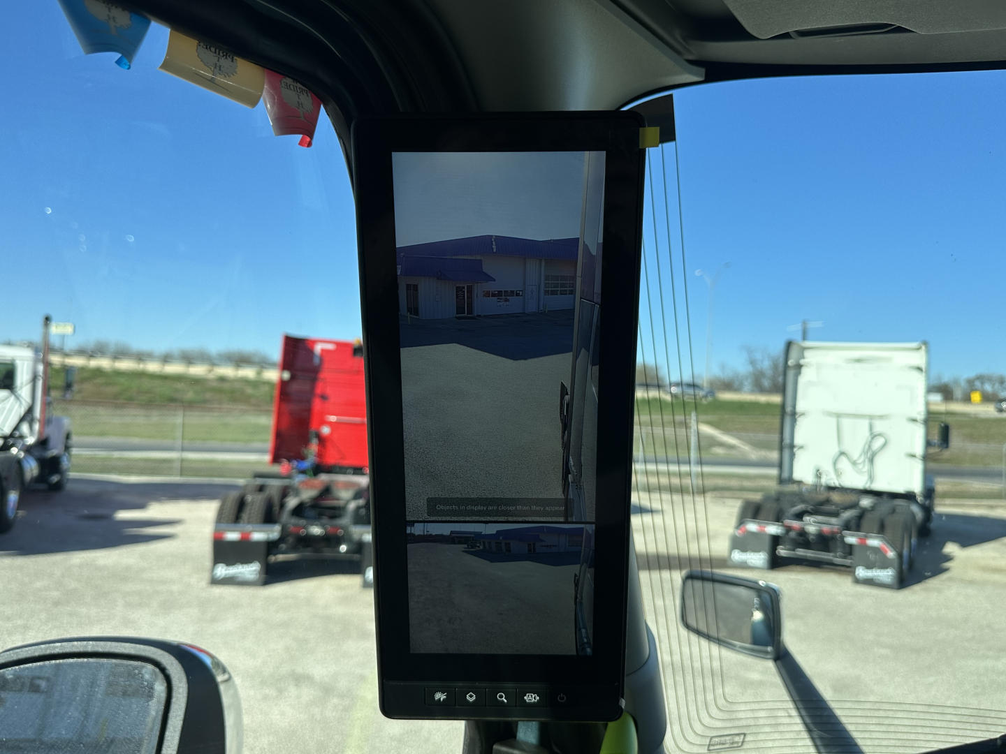 Truck cab interior with a mounted tablet-like display showing outdoor view; window with blue sky; exterior trucks in lot.