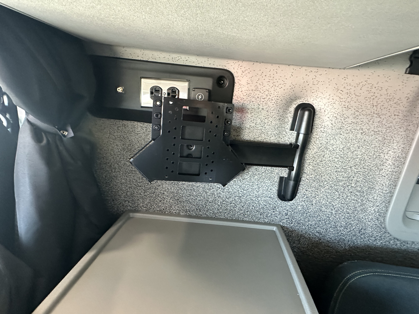 Truck interior showing a wall-mounted monitor/TV bracket and power outlet, with a grey cabinet below and a black handle attached to speckled panel.