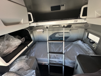 Truck cabin bunk area with ladder and plastic-wrapped mattress; overhead storage pockets visible.