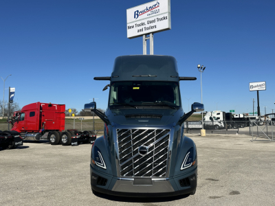 Front view of a new Volvo VNL64T860 truck in a dealership lot, chrome grille, large windshield, side mirrors, and signage in background.
