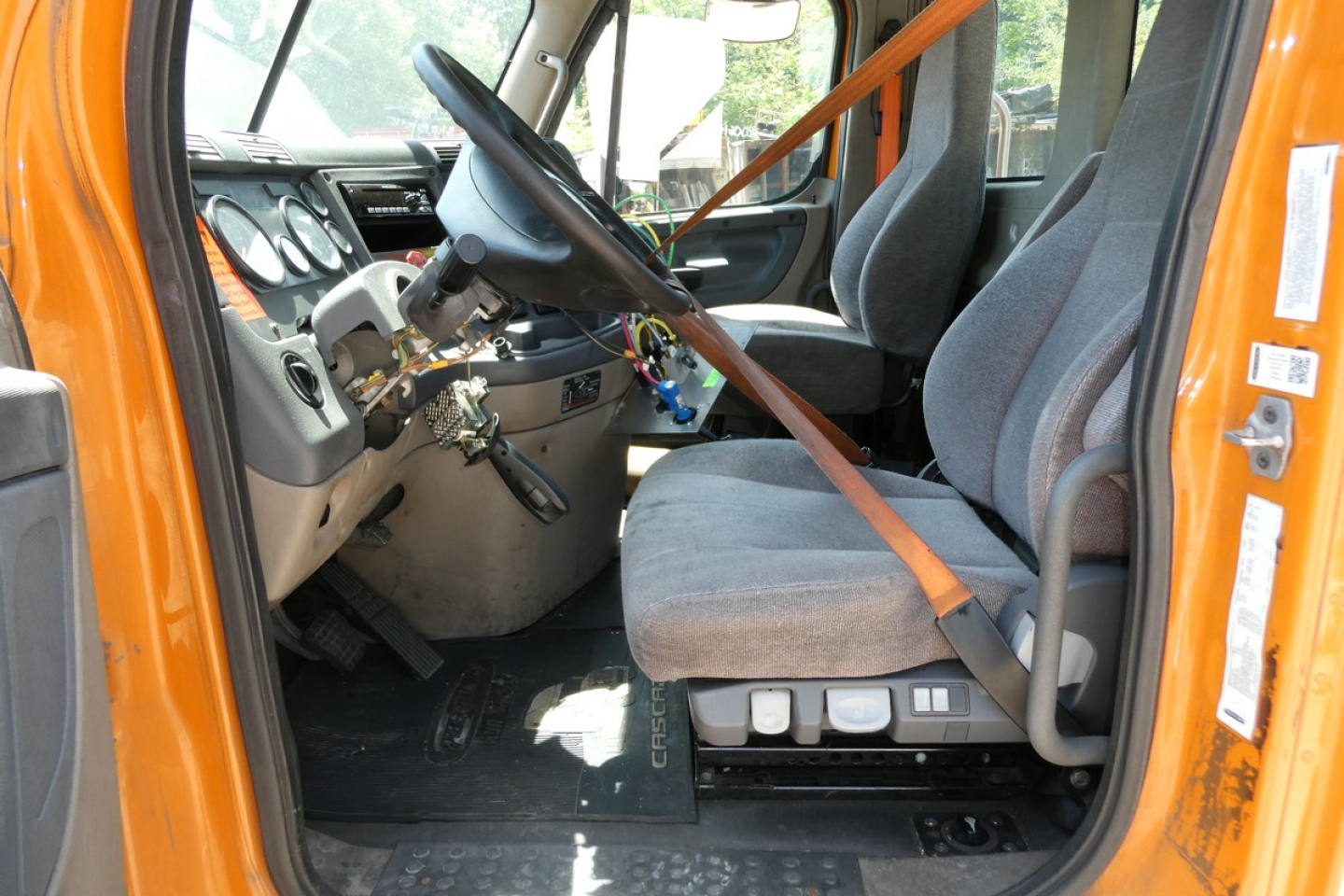 Cab interior of a Freightliner truck showing steering wheel, dashboard, and two gray seats with orange seat belt across. Center console and pedals visible.