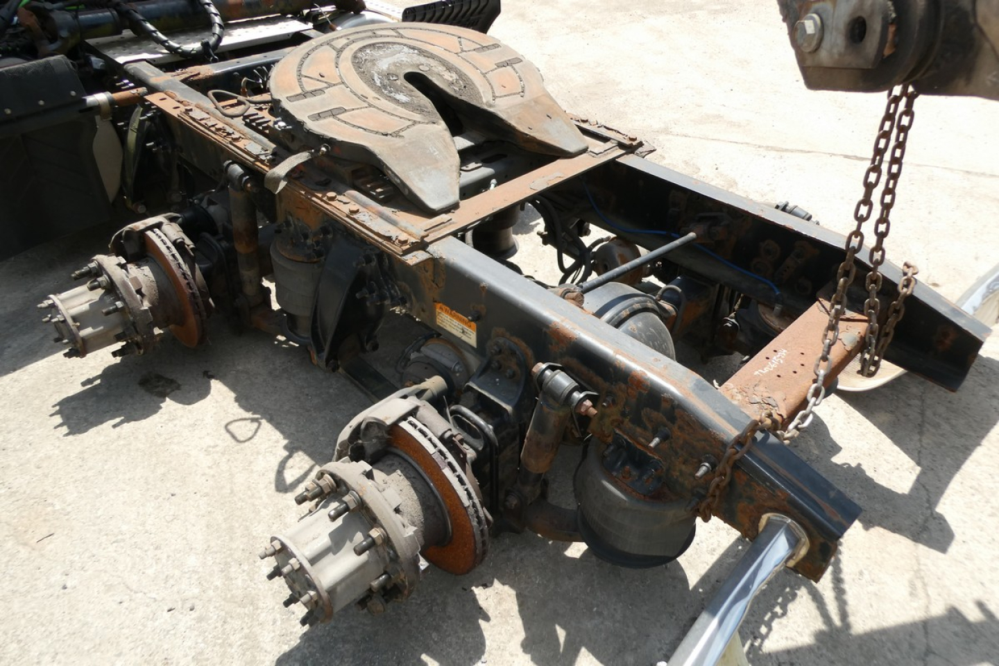 Chassis frame and drive axle components of a Freightliner Cascadia 125 truck without wheels; rusted metal, visible suspension hubs, rotor brake drums, and a chain hanging nearby.
