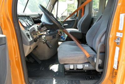 Cab interior of a Freightliner truck showing steering wheel, dashboard, and two gray seats with orange seat belt across. Center console and pedals visible.