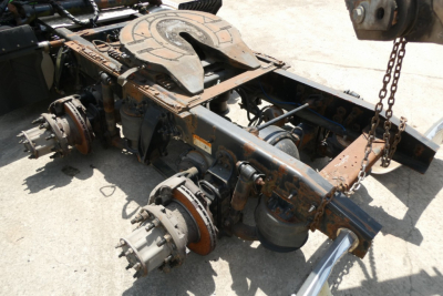 Chassis frame and drive axle components of a Freightliner Cascadia 125 truck without wheels; rusted metal, visible suspension hubs, rotor brake drums, and a chain hanging nearby.