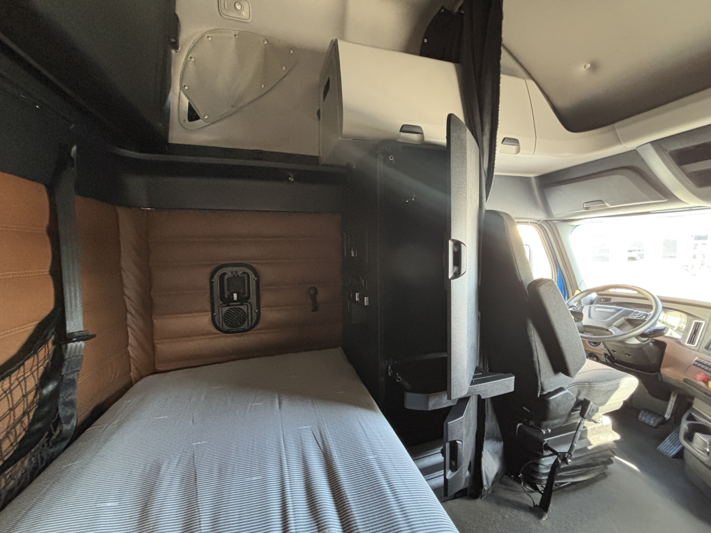 Vehicle cab interior showing sleeper area and driver's seat in a Freightliner truck cab, with a storage box and partition behind the driver.