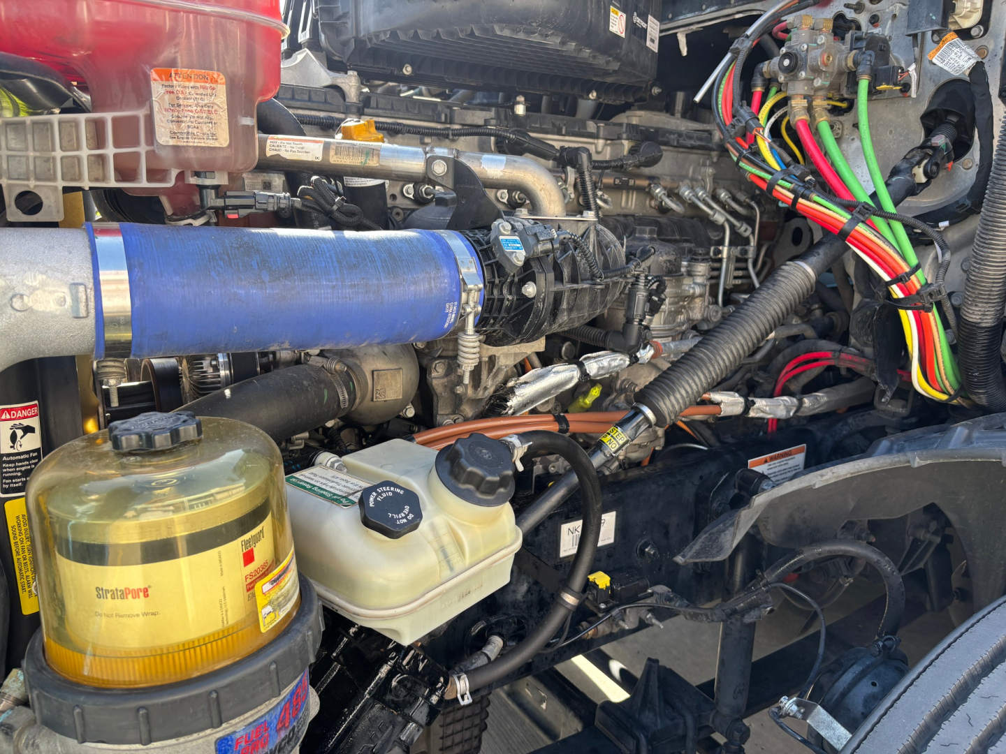 Trucks engine bay with wiring harnesses and blue intercooler hose; coolant reservoir, power steering/washer fluid reservoirs, and various hoses visible.
