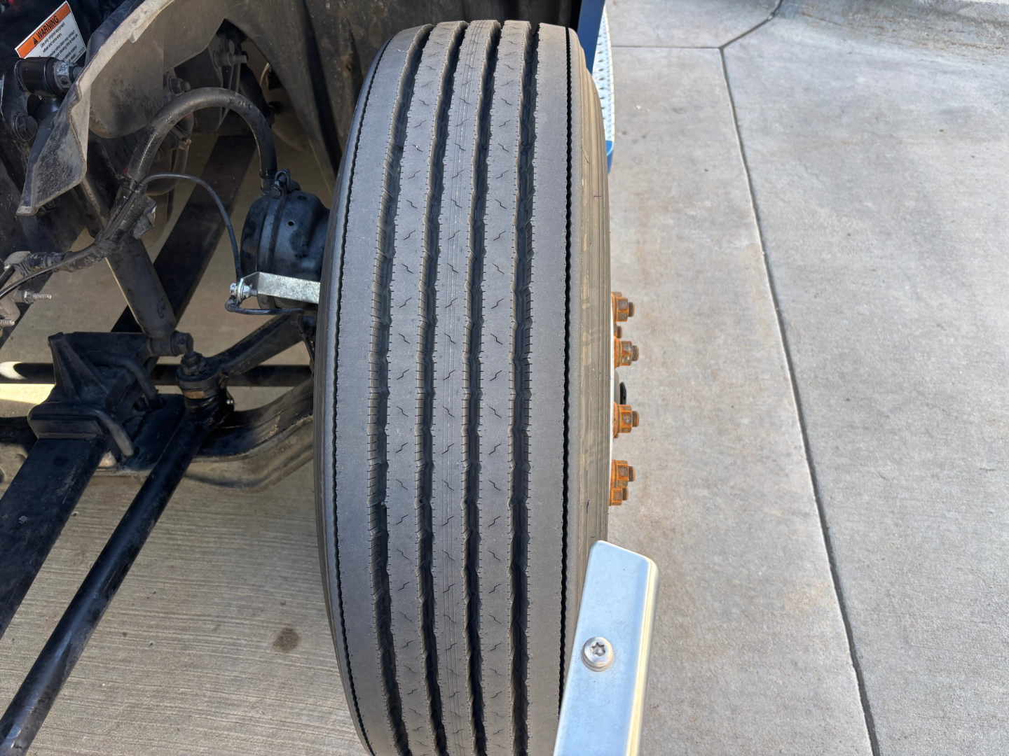 Front wheel view of a 2023 Freightliner PT126064ST conventional sleeper truck showing single tire with dual grooves and adjacent suspension components.
