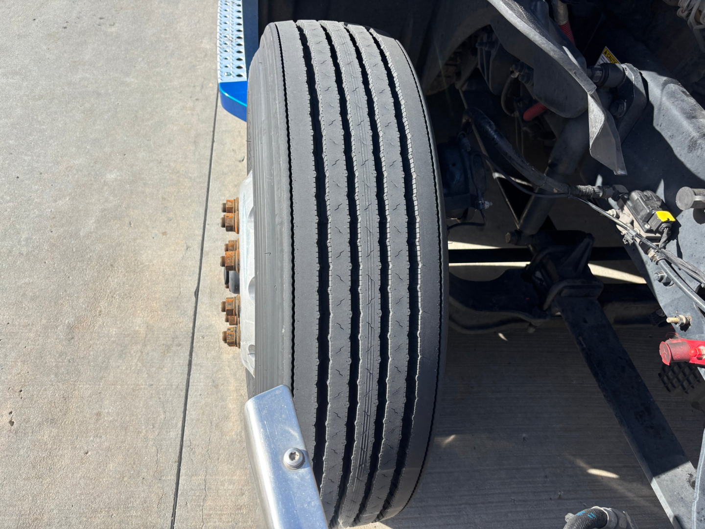 Truck front wheel and suspension view showing dual rear tires and axle components under a Freightliner PT126064ST cab with visible bolts.