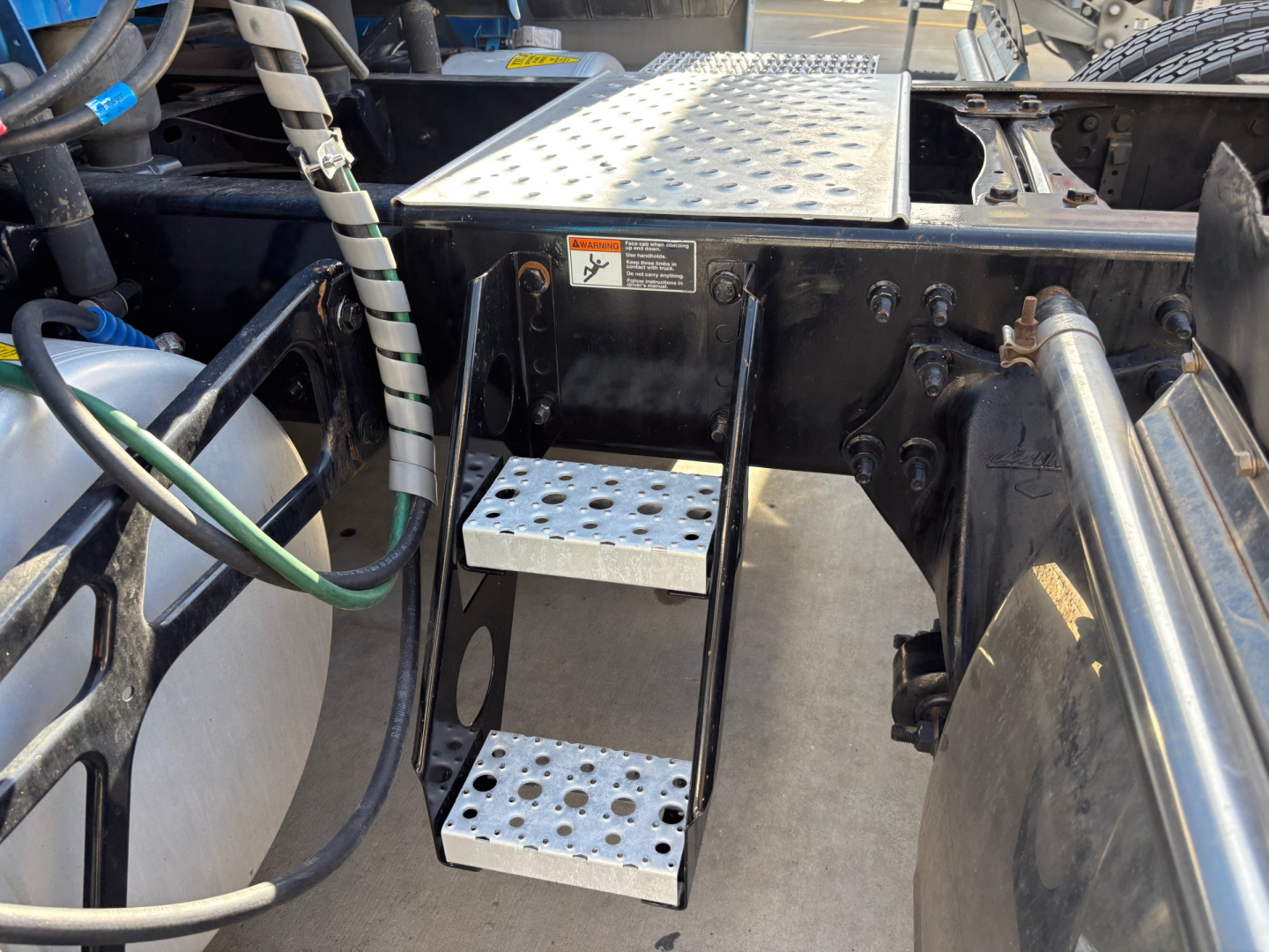 Steps and cab area of a Freightliner truck showing aluminum perforated stepping with footholds and nearby hoses; warning label and metal chassis with diamond plate platform.